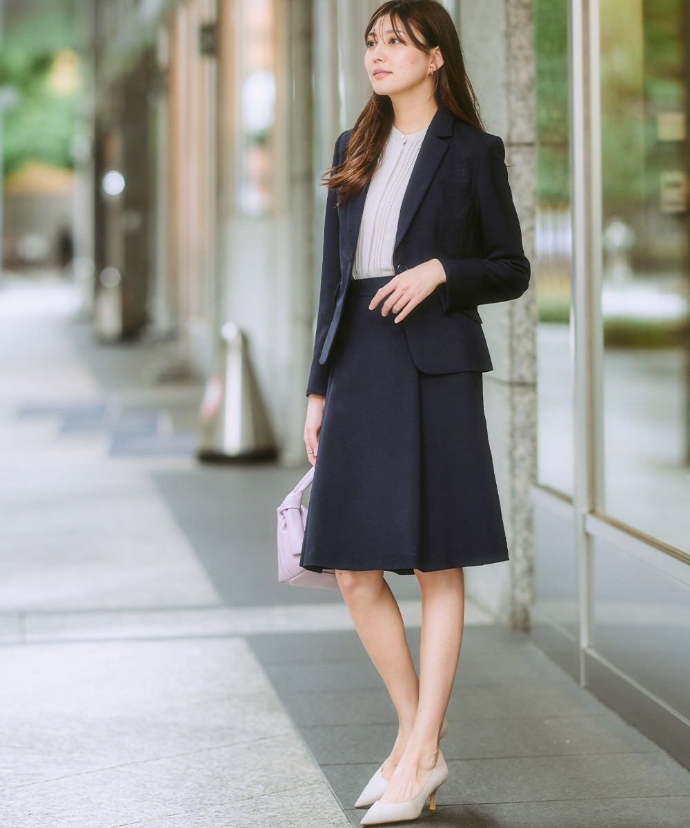 日本女性穿深藍西裝外套、白色細褶上衣、同色打褶 A 字中裙、淺色尖頭跟鞋並提淡紫色手提包
