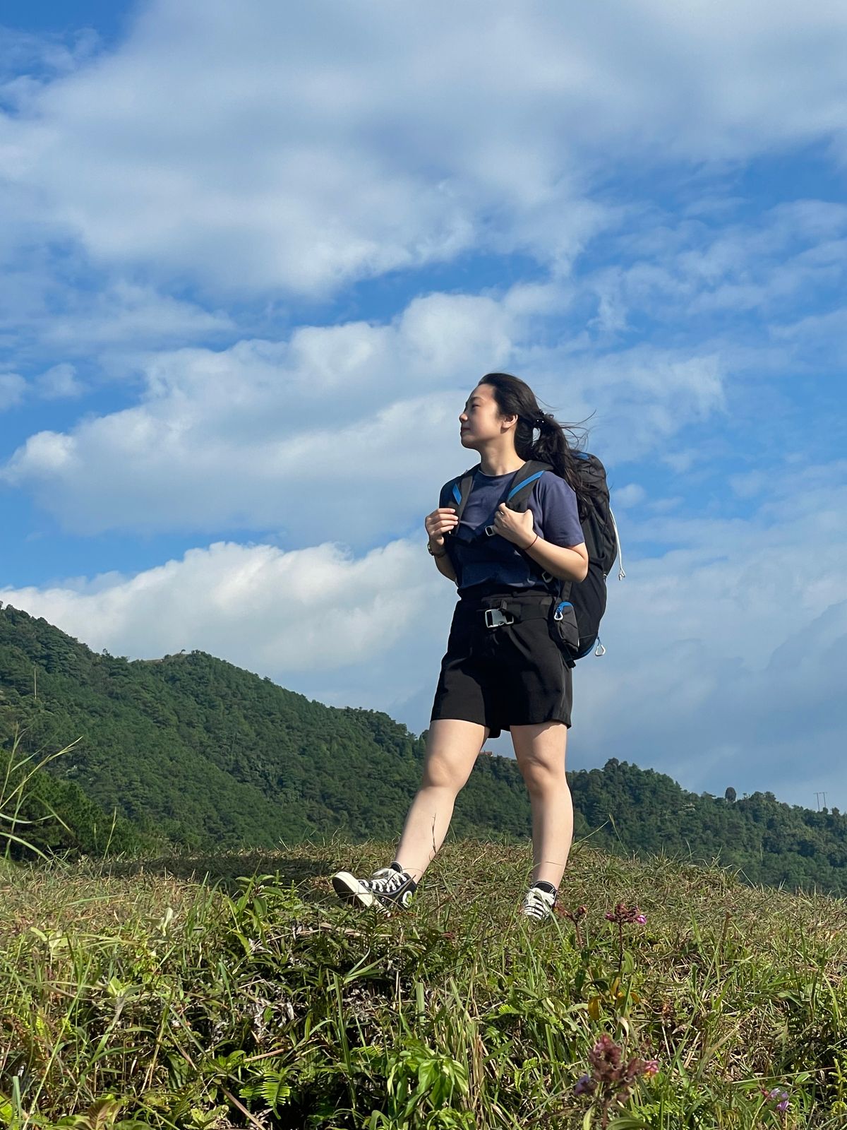 亞洲女性穿深色運動上衣與黑色短褲,背健行背包站在草地山徑