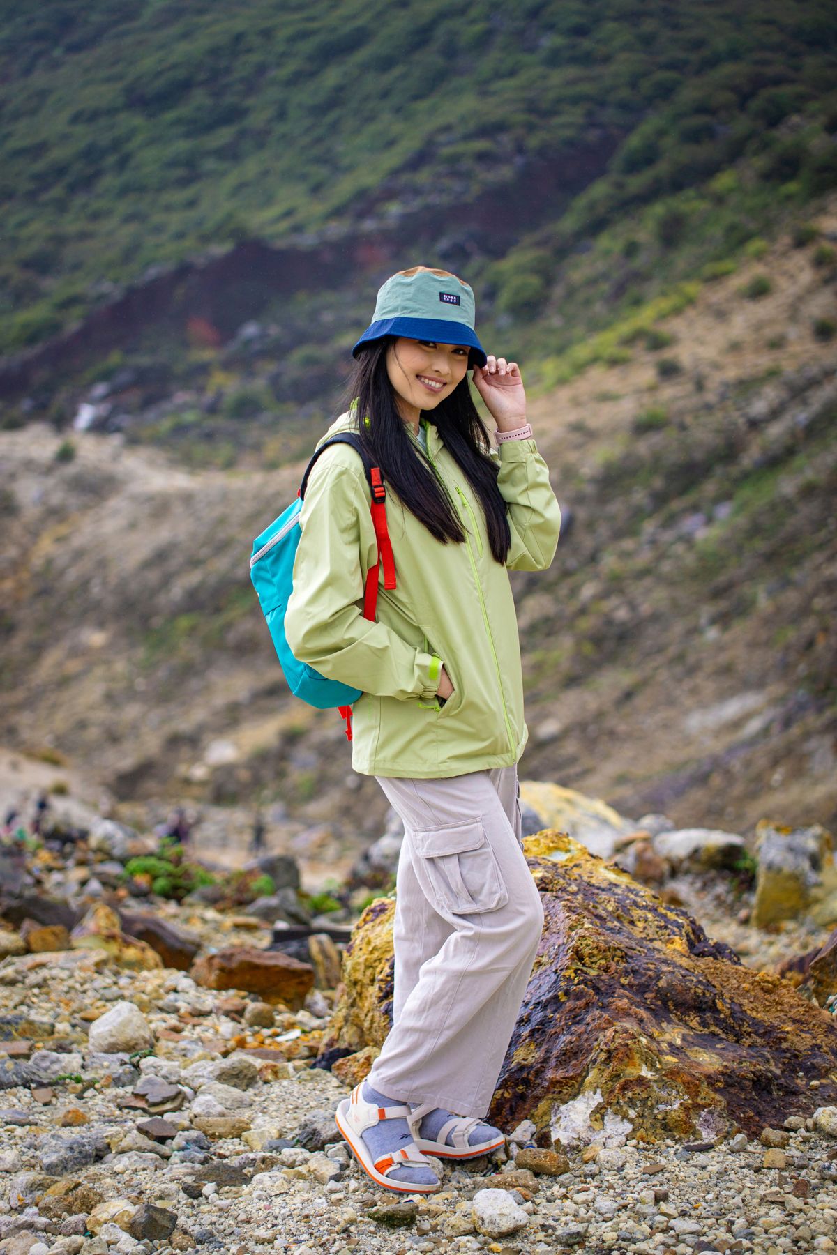 亞洲女性穿綠色戶外外套與淺色工裝褲、戴帽並背彩色背包走山徑