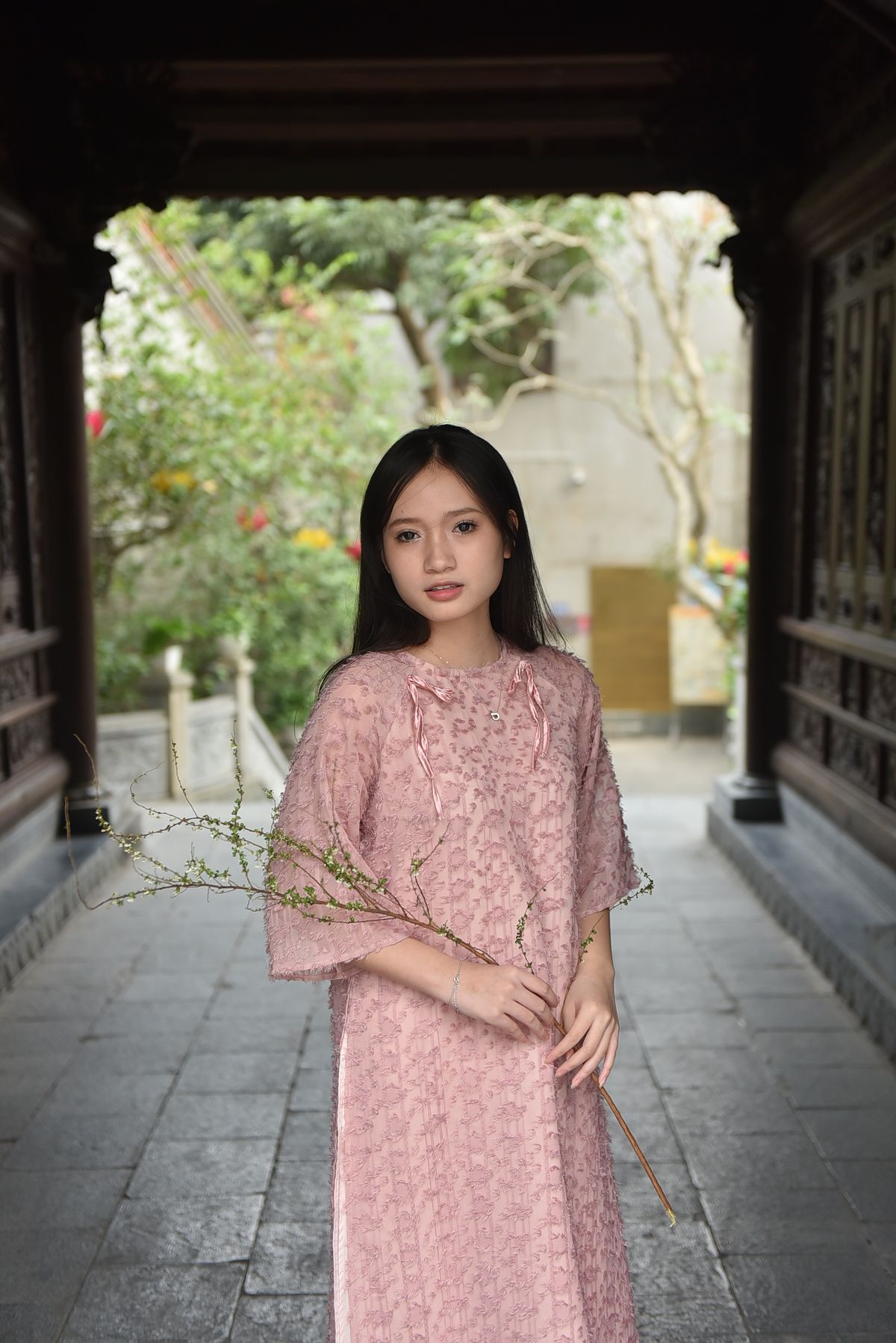 亞洲女性穿粉色小花洋裝,胸前有綁帶細節,站在庭院石廊中