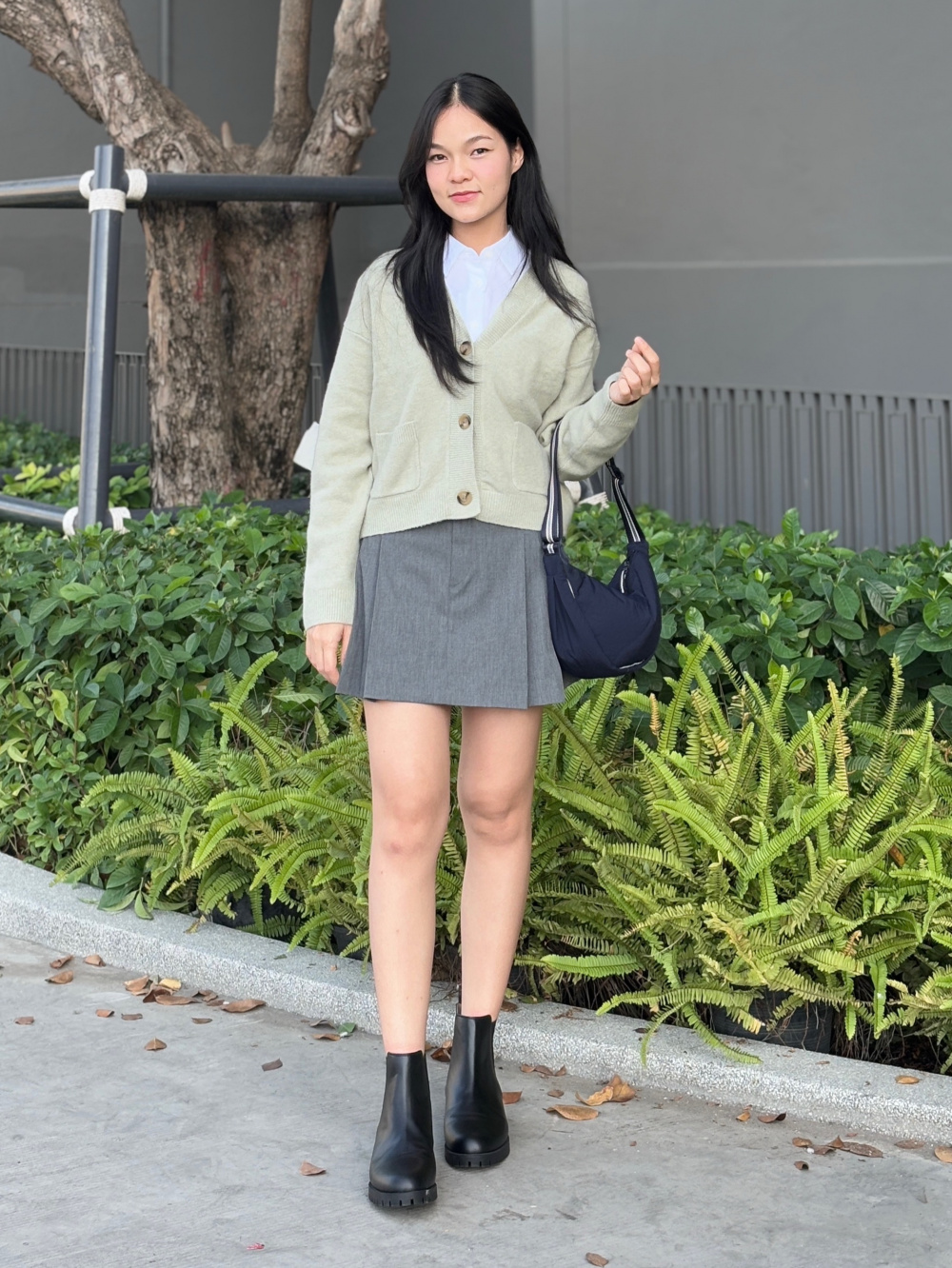 亞洲女性以淺色開襟針織搭白襯衫、灰色短裙、黑色短靴和深色肩背包的全身造型