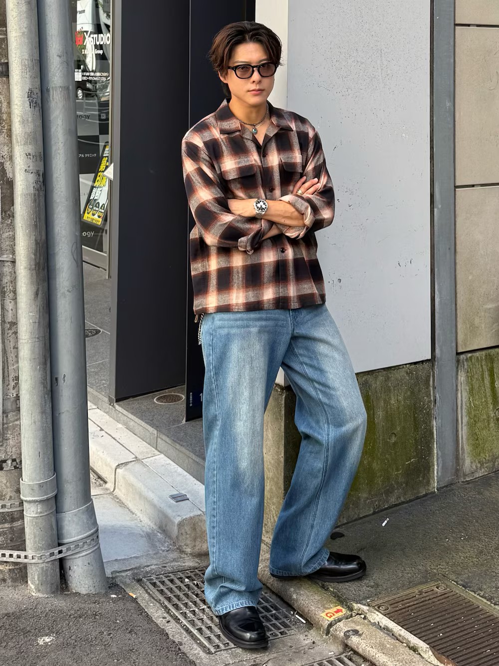 日本男性戴黑色粗框墨鏡,穿棕色格紋襯衫與淺藍寬直筒丹寧,搭配銀色項鍊、手錶與手環